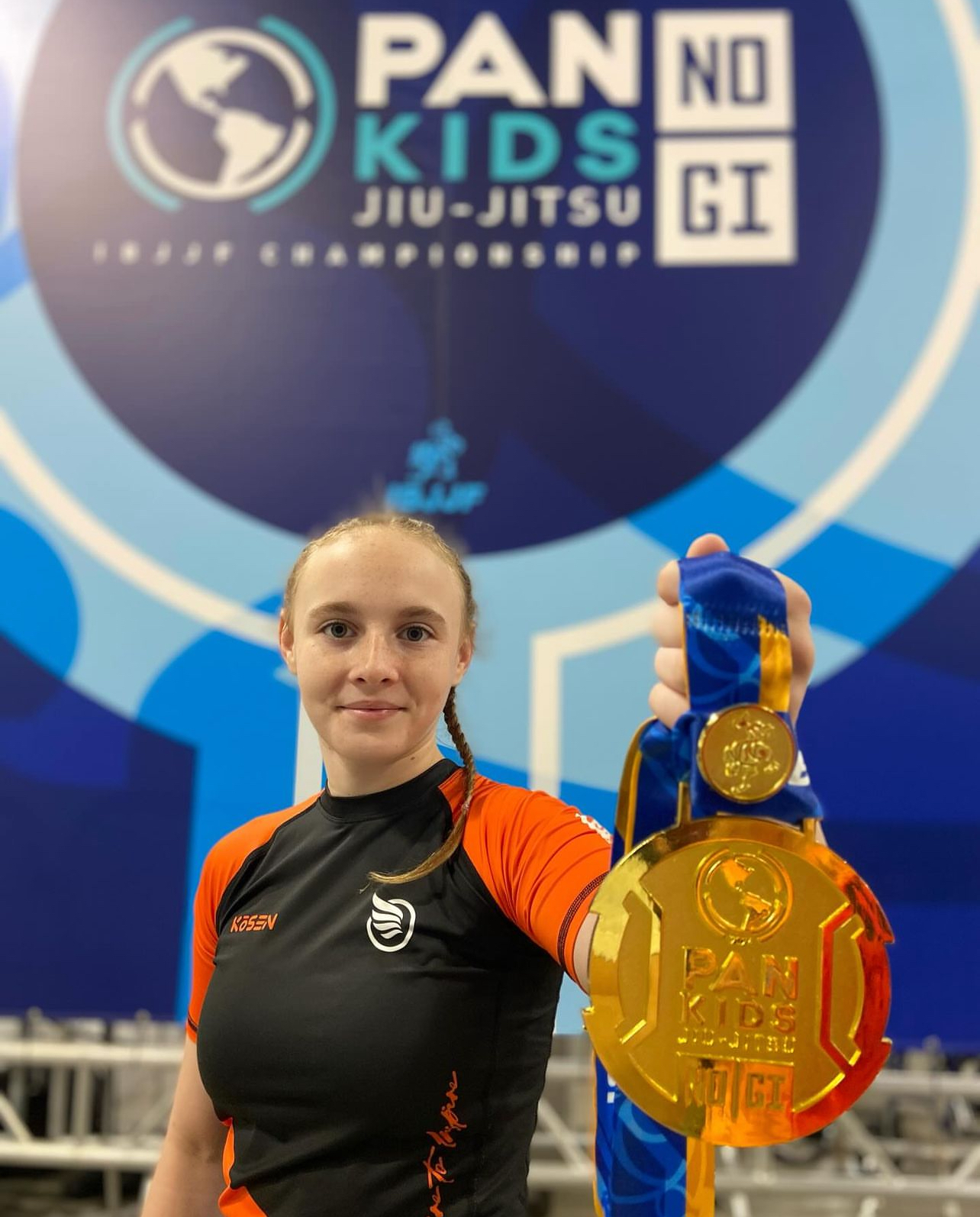 Fotografie der Goldmedaille, aufgenommen bei der IBJJF Pan Kids No-Gi Championship 2024. Die Medaille trägt die Kennzeichnung Pan Kids IBJJF Championship. Im Hintergrund ist die Event-Rückwand PAN KIDS NO-GI IBJJF CHAMPIONSHIP sichtbar.